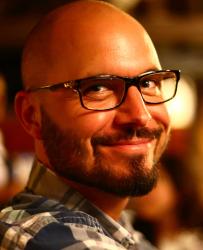 An individual with black glasses and a patterned shirt smiling 